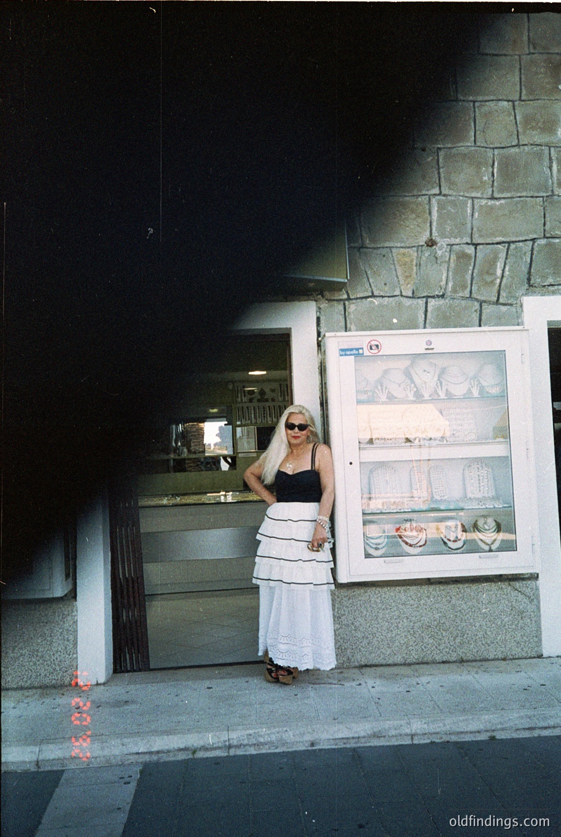 Vintage-style photo of a woman in a 1970s-style swimsuit and sunglasses posing outside a bakery. Concrete building with a glass display showcasing pastries. Sunlight casts shadows on pavement, hinting at Mediterranean or coastal European setting.