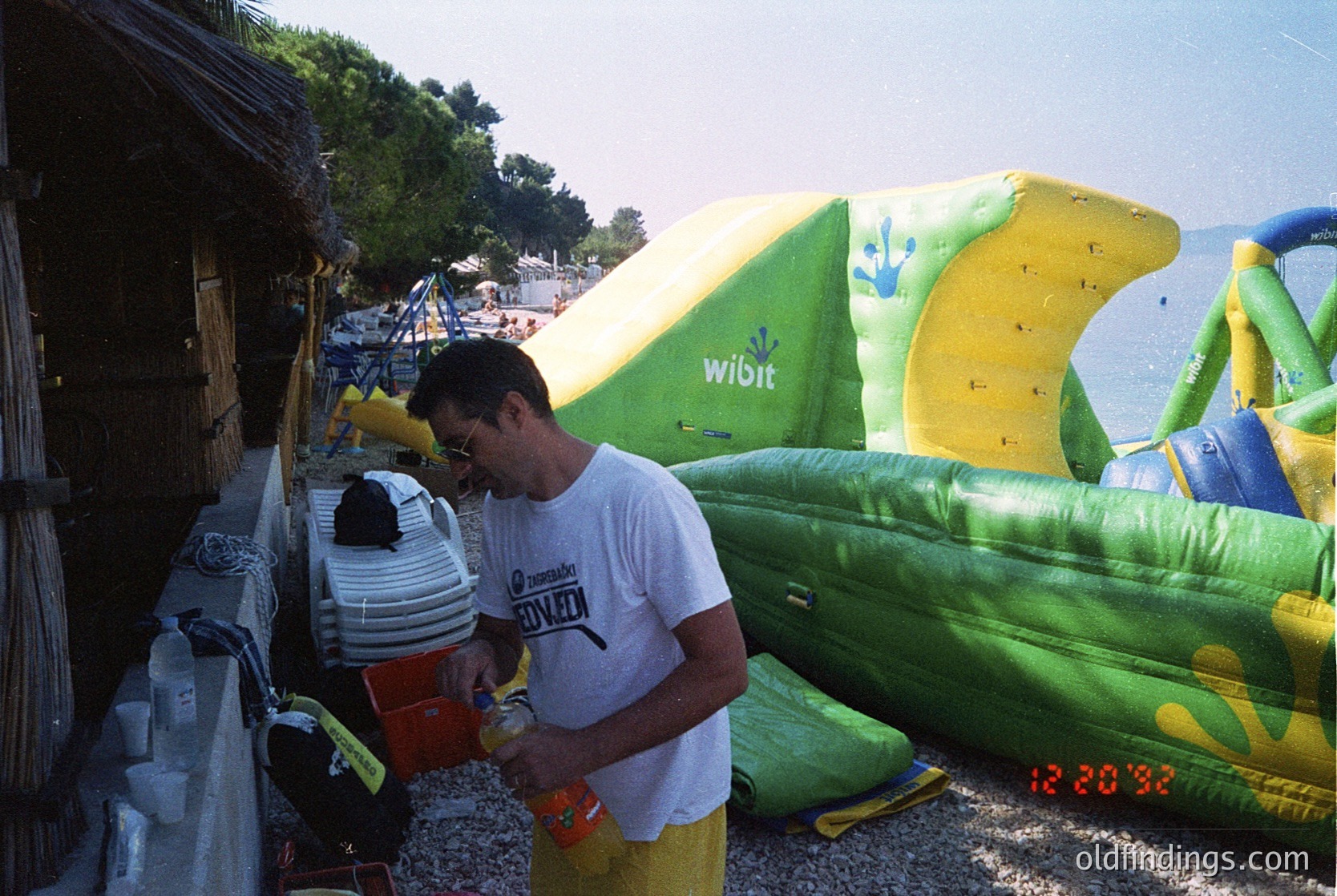 Retro beach scene featuring inflatable water sports equipment. A man in a white T-shirt with a "Wibit" logo on a banana-shaped inflatable and a woman in a striped swimsuit examine a red cooler. Wooden beach huts and a sandy shore with palm trees in the background suggest a Mediterranean coastal resort, likely from the 1990s or early 2000s.