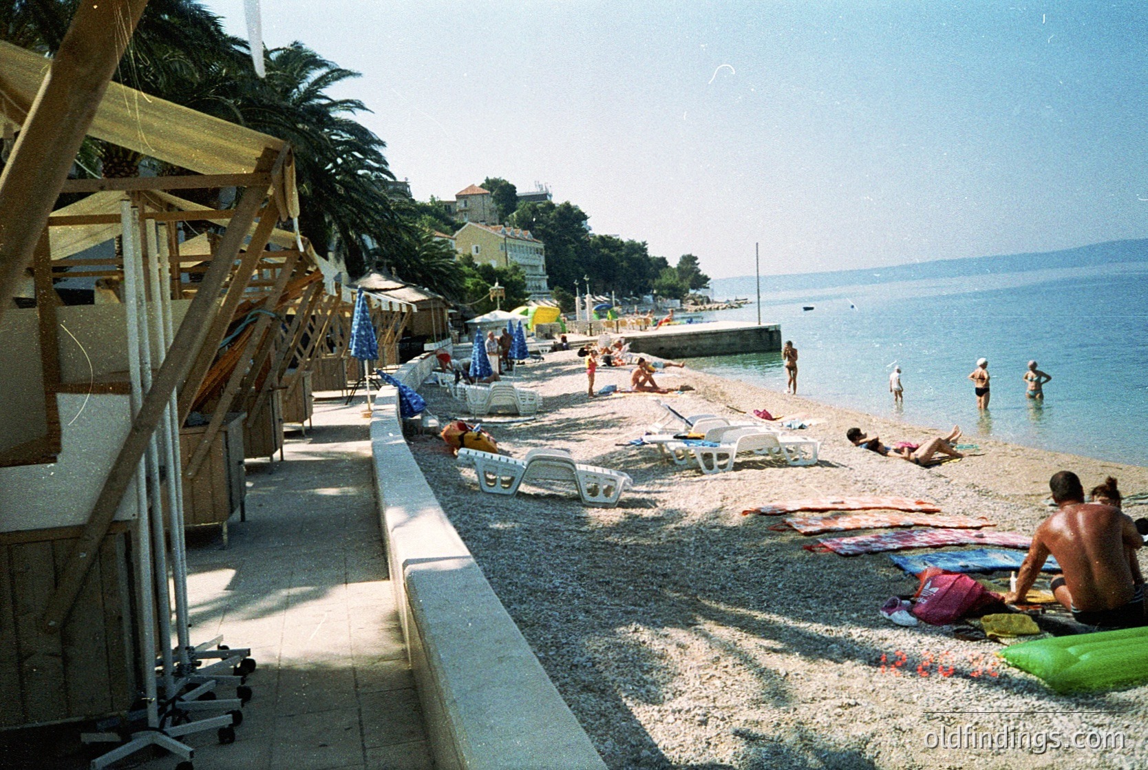 Sun-drenched seaside promenade with mid-20th-century beachgoers lounging on towels and sunbeds. Palm trees line the left, while a concrete pier extends into the turquoise sea. Classic cars parked along the pebble shore. Likely Mediterranean coastal town, 1960s–1970s.