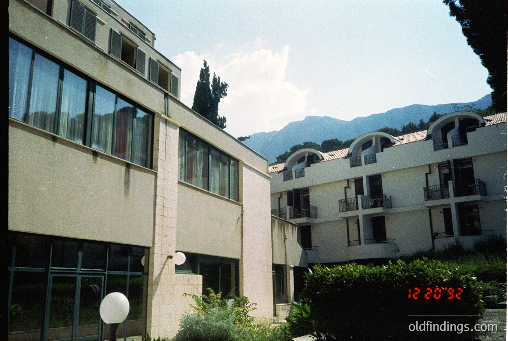 Mid-century modern apartment complex with flat roofs, large windows, and concrete/brick façade. Mountain backdrop suggests alpine or hilly urban setting. Digital timestamp reads 12:20:32.