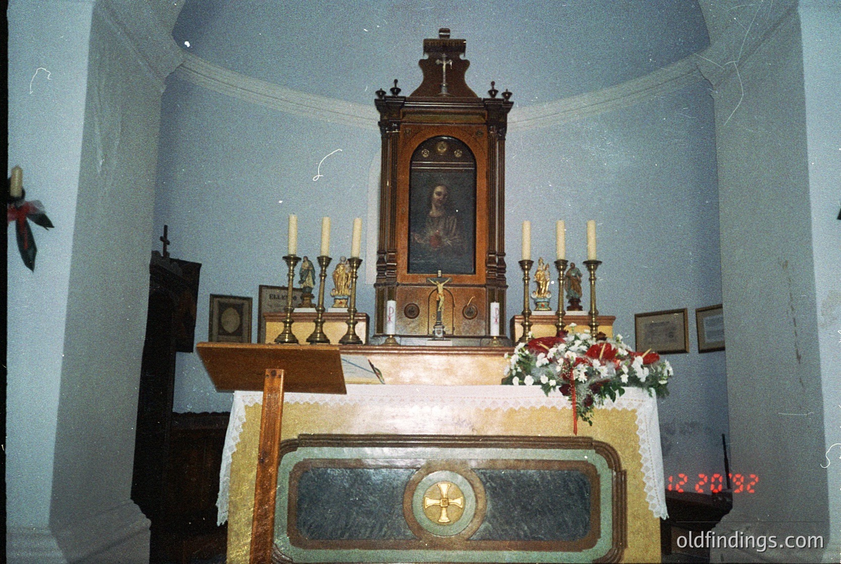 Vintage interior of a religious altar featuring a wooden iconostasis with a framed religious portrait above. Candlesticks, a cross, and floral arrangements adorn the altar table. Architectural details include arched ceilings and a digital clock (12:20:32) in the lower right corner.