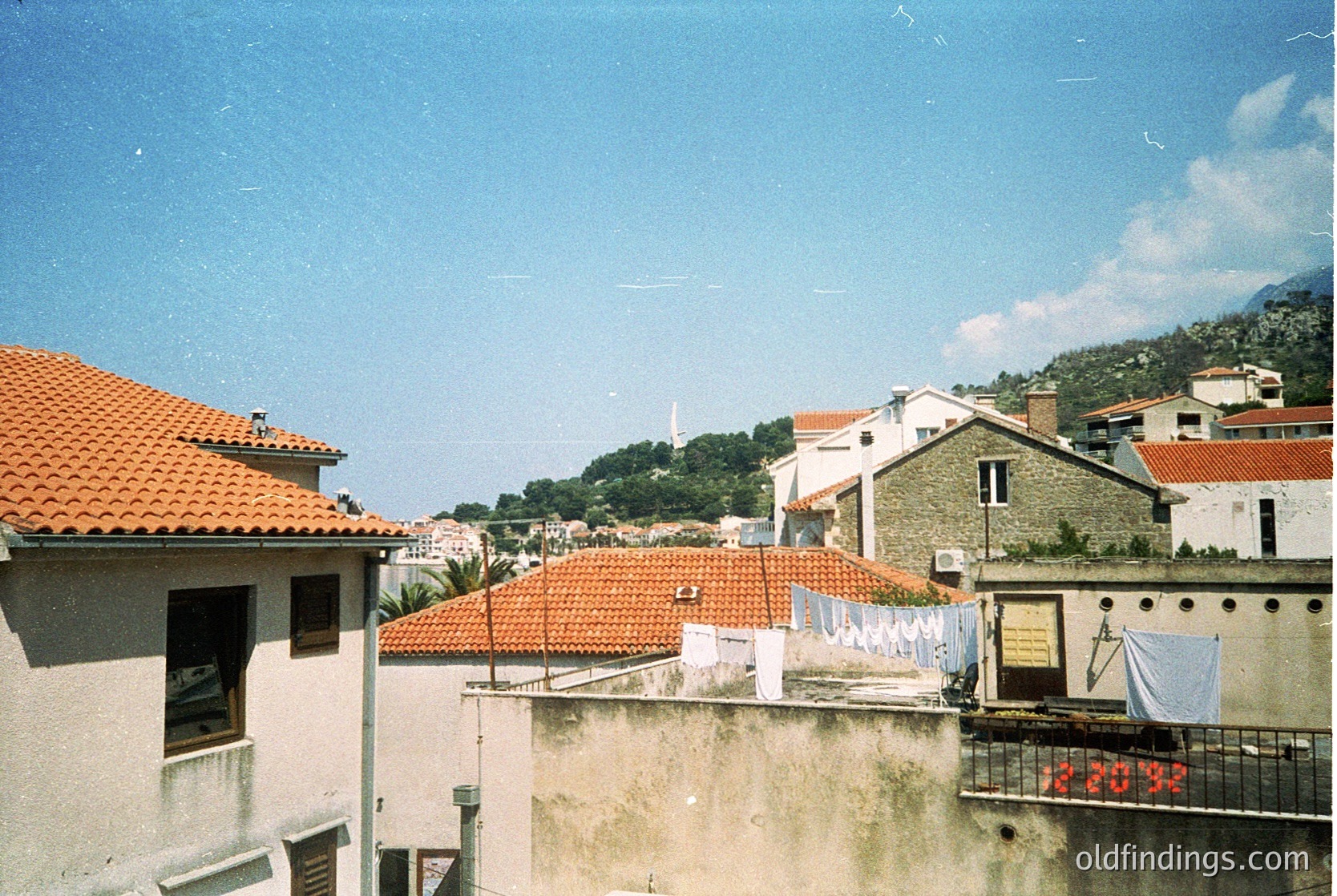 Vintage aerial view of Mediterranean hillside village with terracotta-tiled rooftops and whitewashed buildings. Laundry hangs on lines, suggesting residential daily life. Lush greenery and distant mountains frame the scene. Likely or Mediterranean architecture.