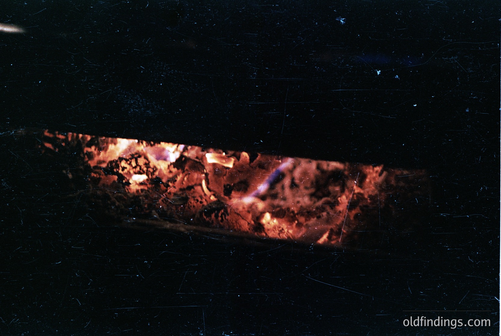 Intense close-up of a burning log fire in a dark setting, showcasing glowing embers and flickering flames. The scene captures raw, warm hues against a stark black background, emphasizing texture and light contrast.