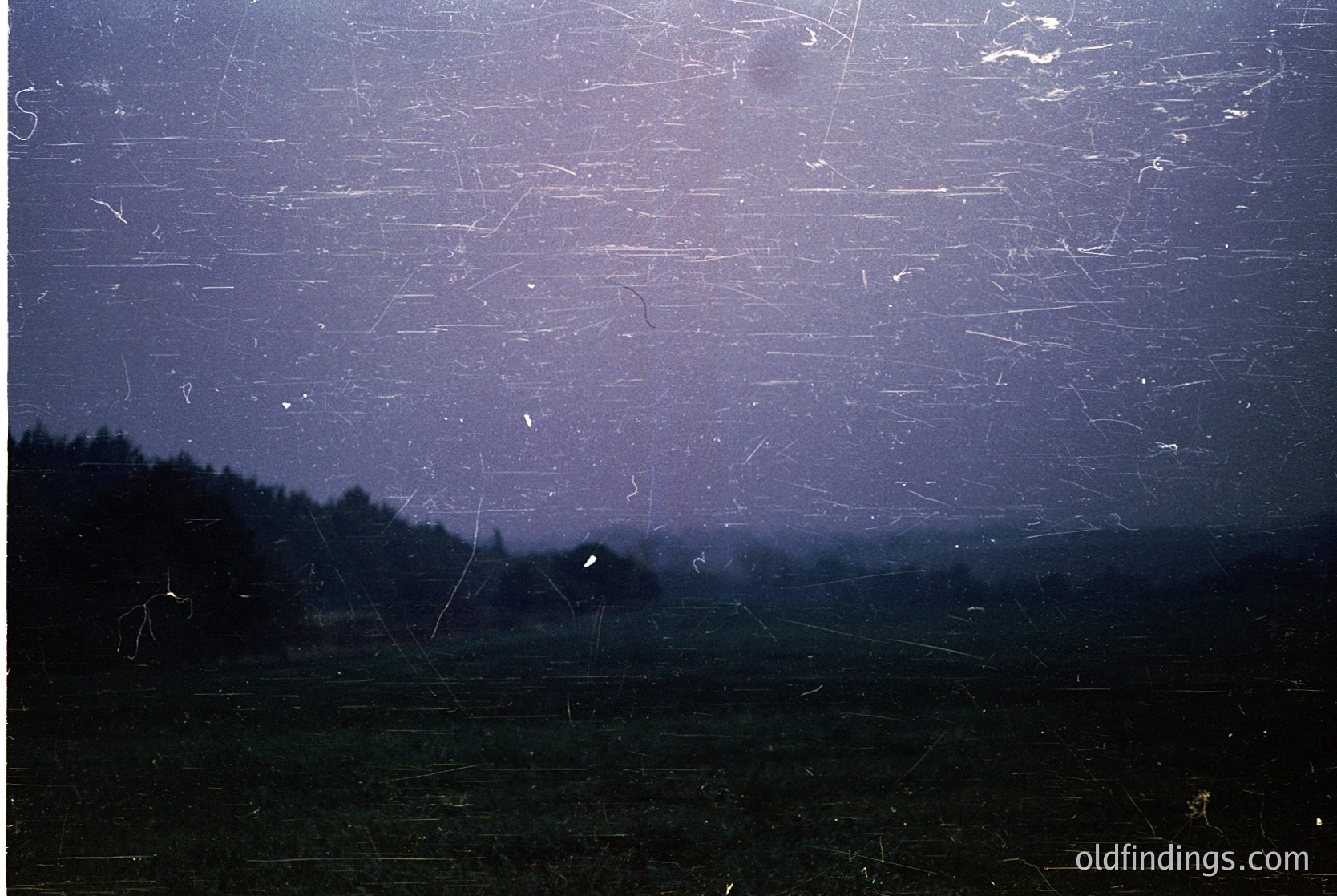 Vintage film grain overlay reveals a moody, rural landscape under storm clouds. Dark horizon silhouettes dense forest and distant fields. Light streaks suggest motion blur from passing rain or camera shake. Likely 20th-century analog photography ().