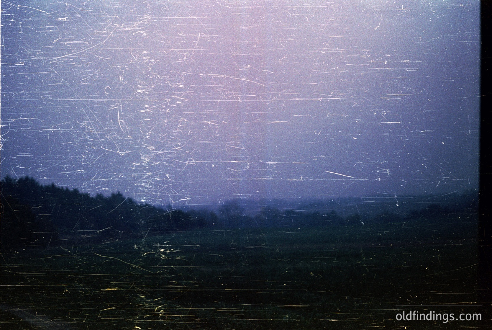 Vintage film grain overlay obscures a hazy, muted landscape with faint horizontal lines suggesting industrial or agricultural structures. Distant silhouettes of trees and fields hint at rural scenery. Likely mid-20th century analog photography ( ).