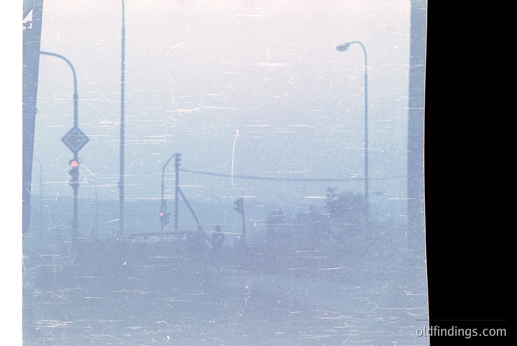 Urban street scene obscured by heavy fog, featuring blurred streetlights, traffic signals, and a pedestrian crossing sign. The muted tones and low visibility suggest early morning or industrial haze. Likely 20th-century cityscape, possibly or .