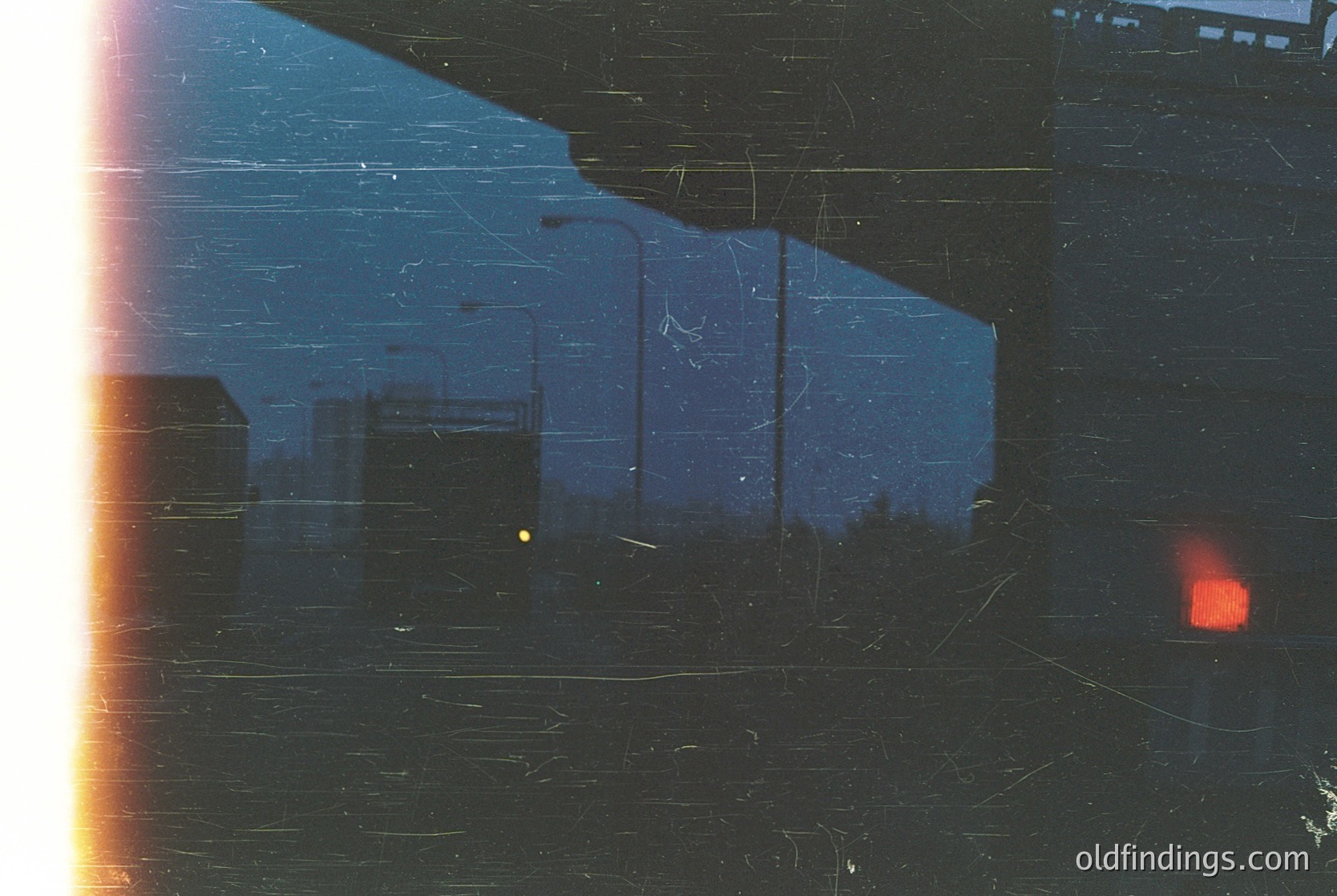 Vintage urban scene viewed through scratched, aged glass—blurred industrial buildings, faint streetlight glow, and a lone red light source. Likely 20th-century cityscape, possibly European.