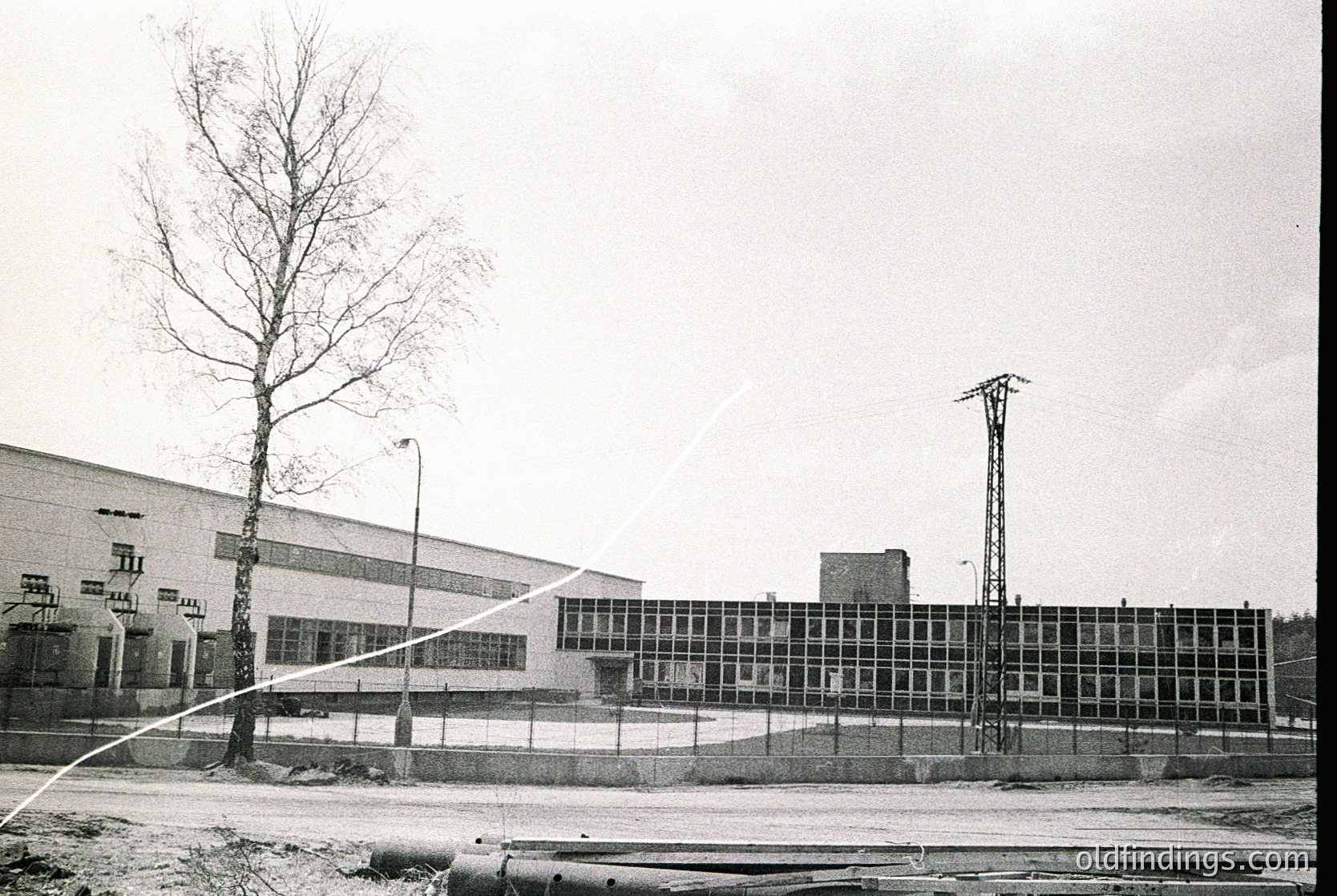 Industrial building with large glass-paneled facade, likely a factory or warehouse, framed by a chain-link fence. Bare tree and utility tower in foreground. Mid-20th century Soviet-era architecture. Overcast sky suggests cold climate.