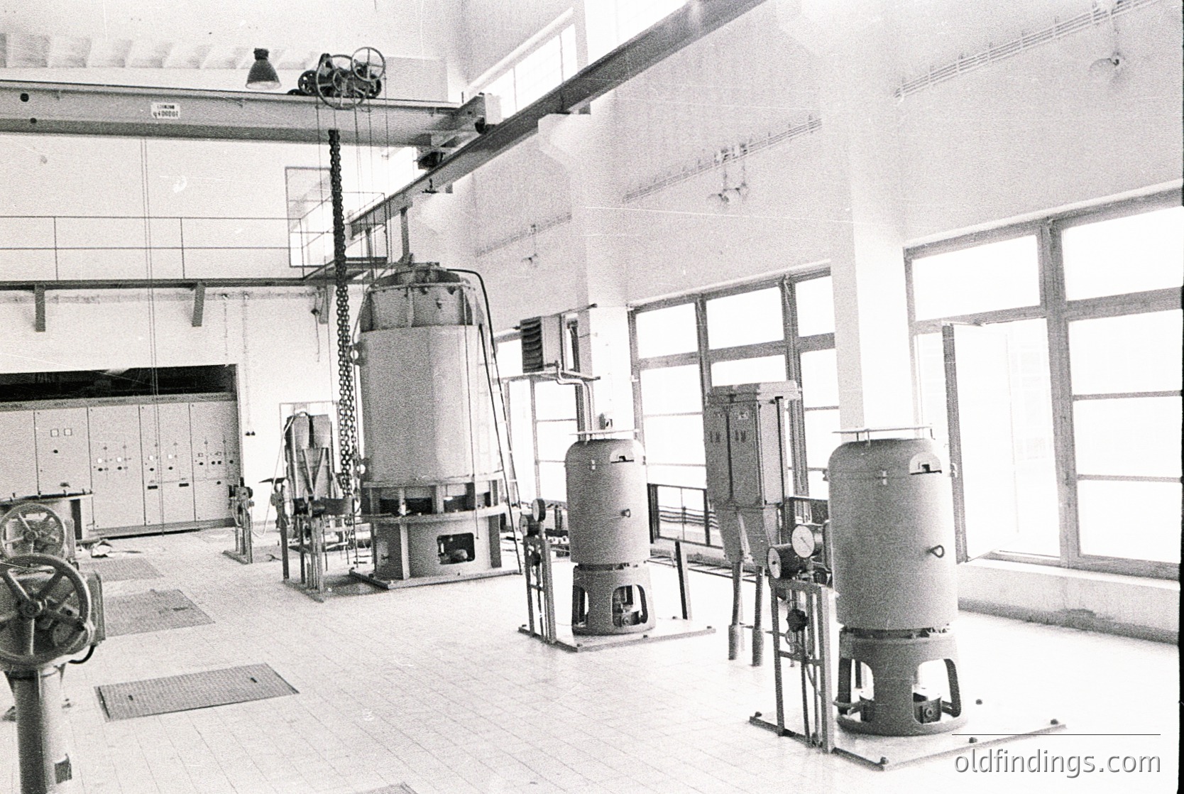 Industrial facility featuring mid-20th century machinery: large cylindrical pressure vessels, overhead conveyor system, and electrical panels. Clean, utilitarian architecture with high ceilings and large windows. Likely a manufacturing or processing plant, possibly from the 1950s–1970s.