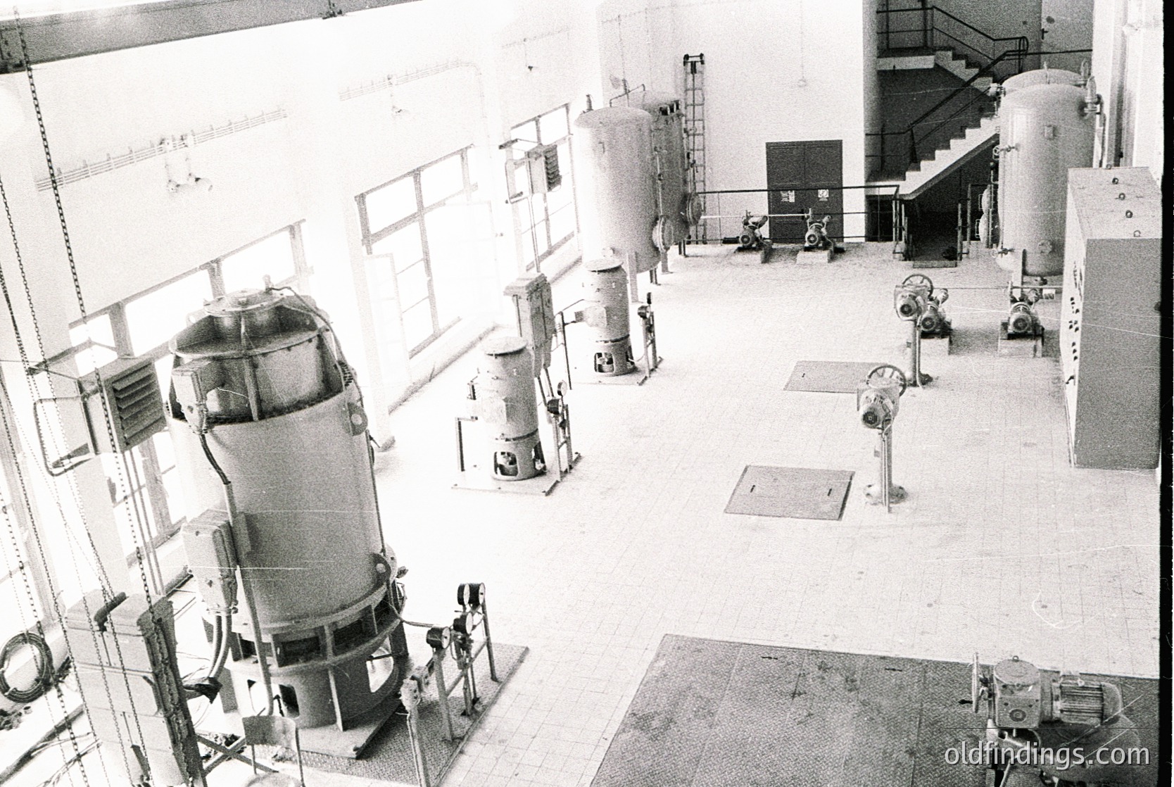 Industrial facility featuring mid-20th century machinery: large cylindrical tanks, valves, and piping in a spacious, concrete-floored room with high ceilings. Staircase and ladder access to upper levels. Likely a chemical, pharmaceutical, or food processing plant.