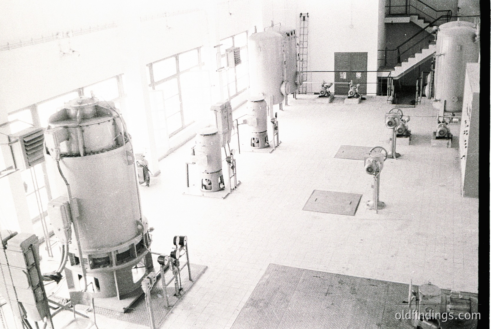 Industrial facility featuring multiple cylindrical tanks and mechanical equipment, likely a dairy or beverage processing plant. Mid-20th century design with exposed piping, staircases, and ventilation fans. Clean, utilitarian architecture with large windows for natural light.
