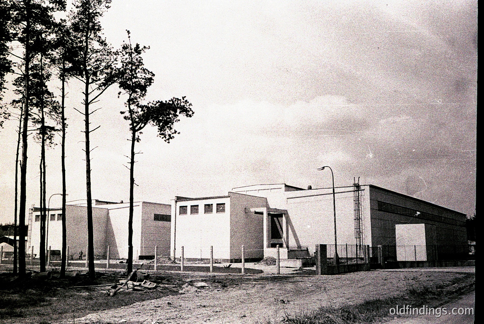 Mid-century modern industrial building with clean lines, flat roof, and large garage doors, surrounded by young pine trees. Concrete and brick construction with minimalist geometric windows. Likely Eastern Bloc-era architecture, 1960s-1970s.