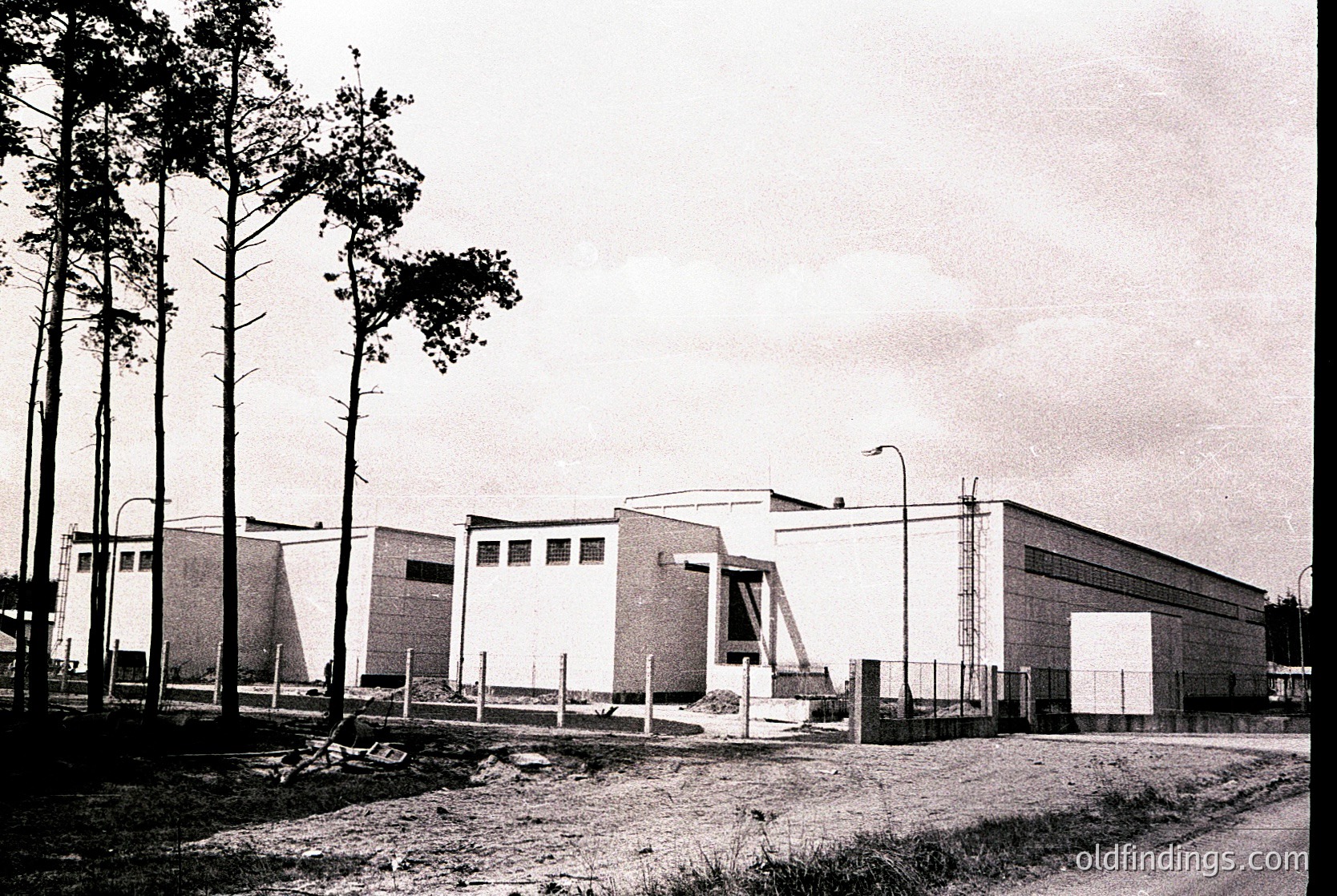 Mid-century modern industrial building with clean lines, flat roof, and large rectangular windows. Surrounded by tall pine trees and a gated perimeter. Likely a warehouse or factory, reflecting 1950s-1960s Brutalist or functionalist architecture.