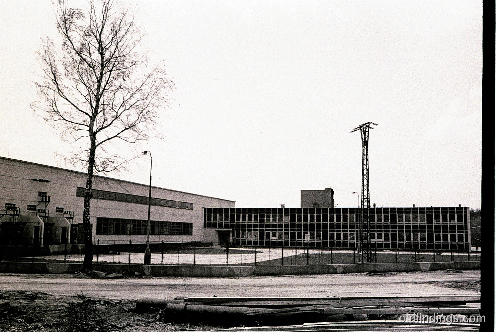 Mid-century industrial building with Brutalist concrete façade and large grid windows, flanked by a lone tree and railway tracks. Likely 1960s–1970s construction.