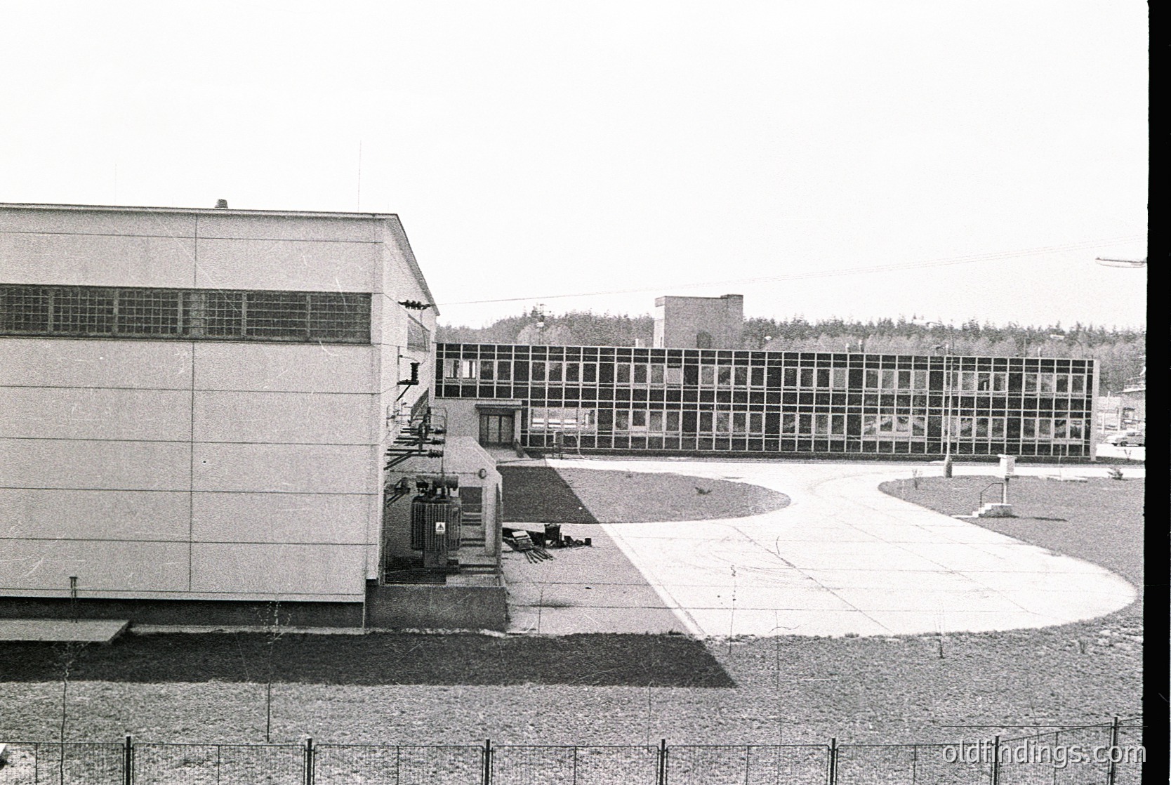 Mid-century Brutalist institutional building complex with raw concrete facade and extensive window bands. Courtyard area with paved surfaces and minimal landscaping. Likely educational or governmental use, 1960s-1970s era.