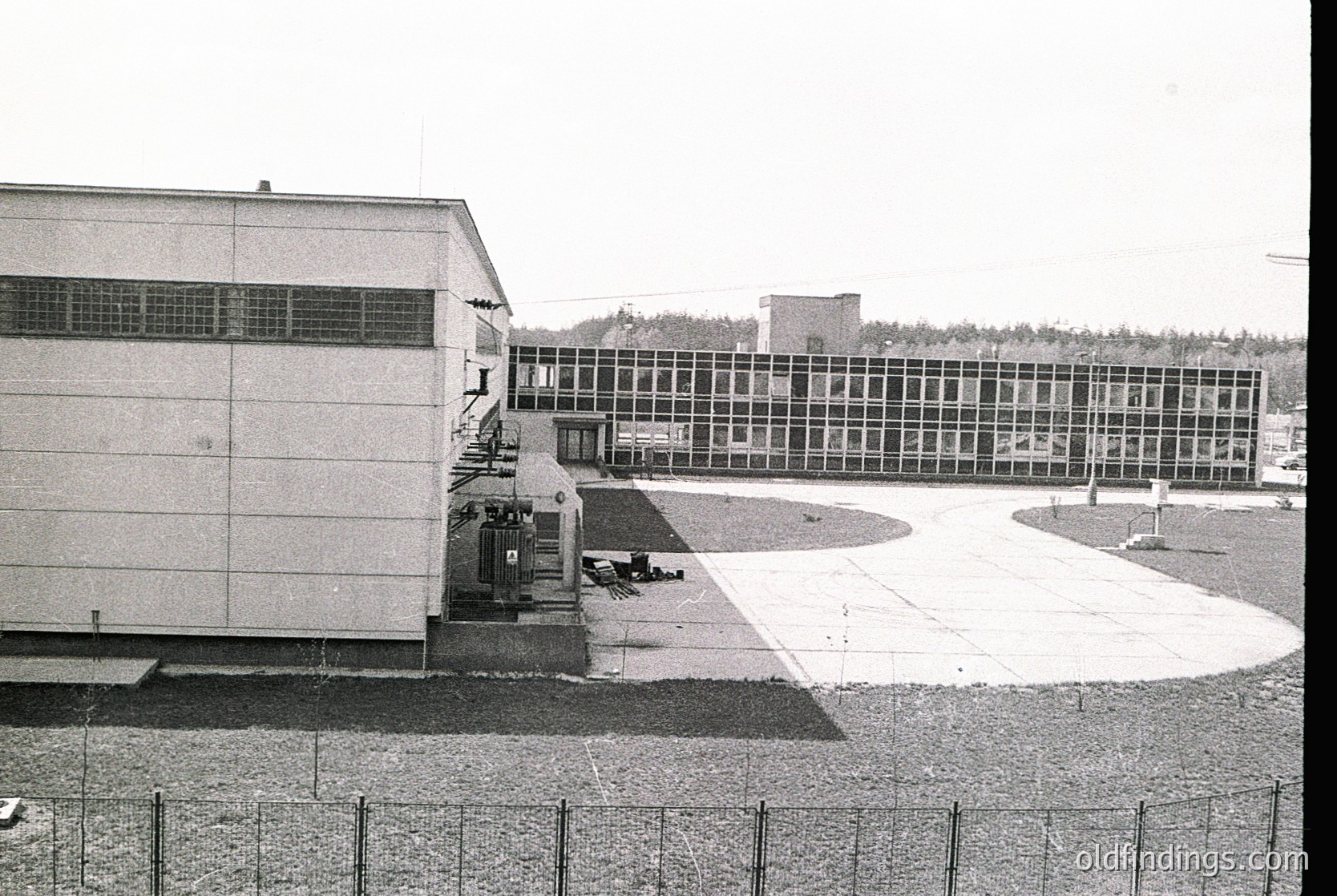 Mid-century Brutalist-style institutional building complex with raw concrete façade and grid-patterned windows. Courtyard area with minimal landscaping, featuring a central paved space and circular concrete elements. Likely a school or government facility from the 1960s–1970s.