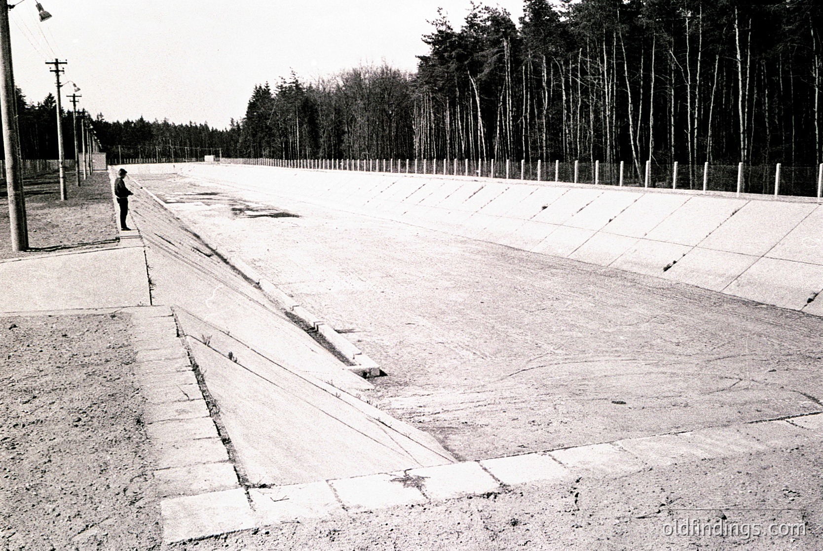 Concrete platform with deep grooves, flanked by barbed-wire fencing and young trees. A lone figure stands near the entrance, suggesting restricted access. Likely a memorial or former detention site, mid-20th century.