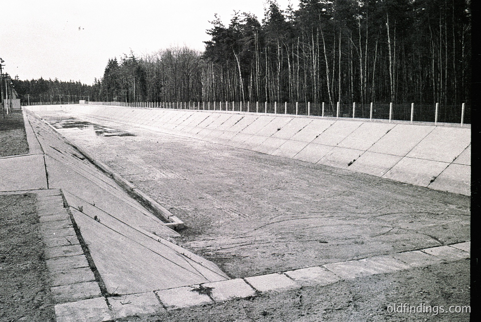 Empty concrete pathway flanked by barbed-wire-topped fences, flanked by dense forest. Likely a WWII-era concentration camp or forced labor site. Structural wear suggests mid-to-late 20th century.