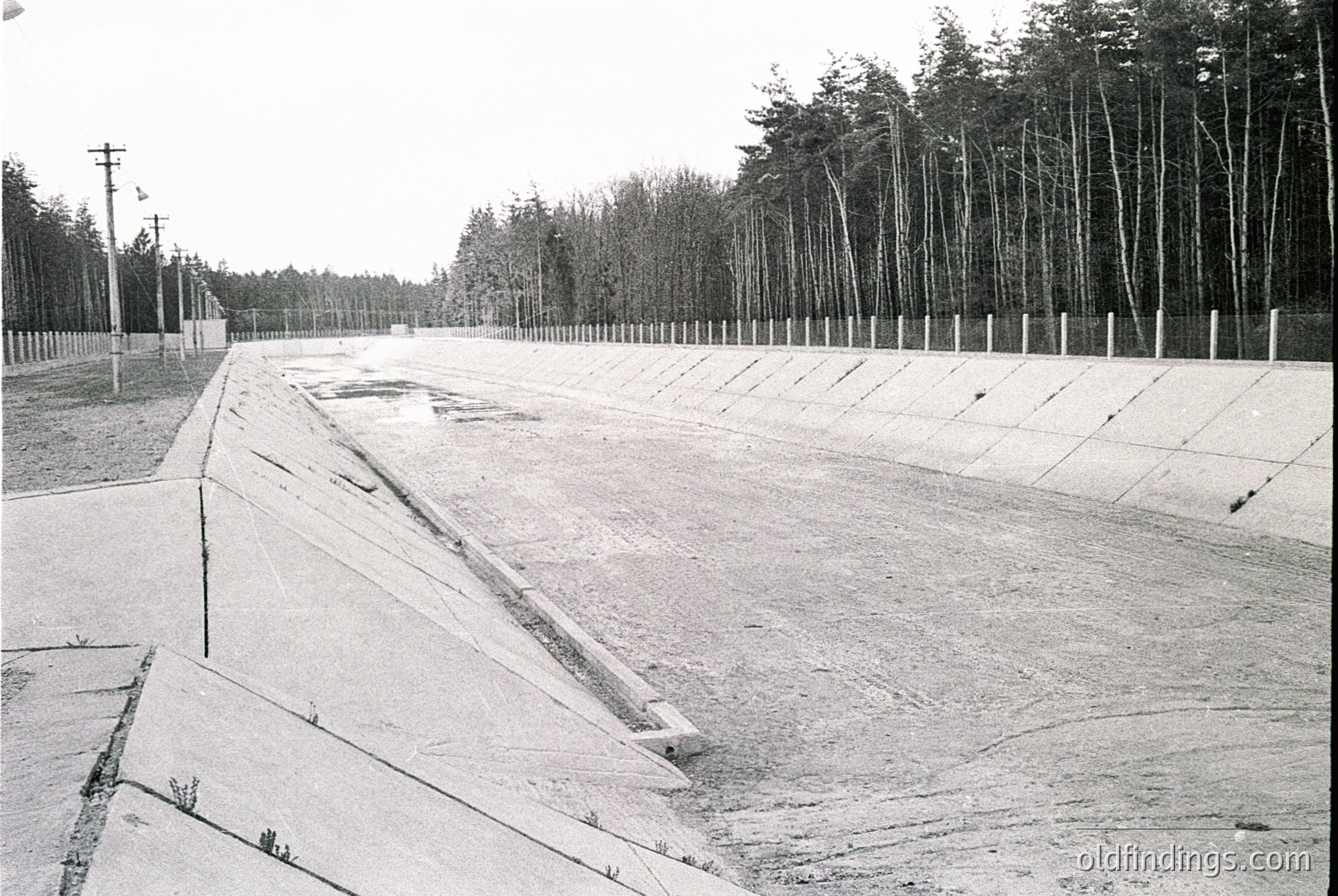 Empty concrete stadium with tiered seating, likely mid-20th century. Rows of wooden poles line the perimeter, possibly for lighting or fencing. Dense forest backdrop suggests rural or suburban location. Snow covers ground, indicating winter season.