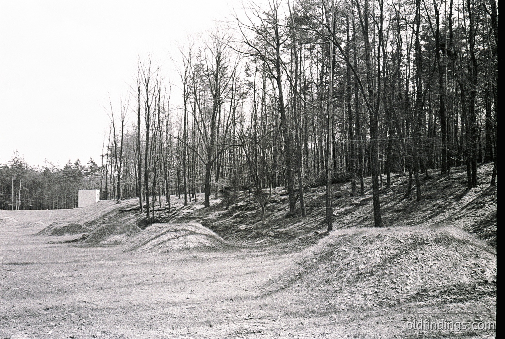 Barren, snow-covered field bordered by leafless trees, likely late winter/early spring. Distinctive earthen berms or ridges suggest agricultural terracing or military fortifications. Minimal structures visible, including a small rectangular building in the background. Black-and-white, suggesting vintage or archival quality.