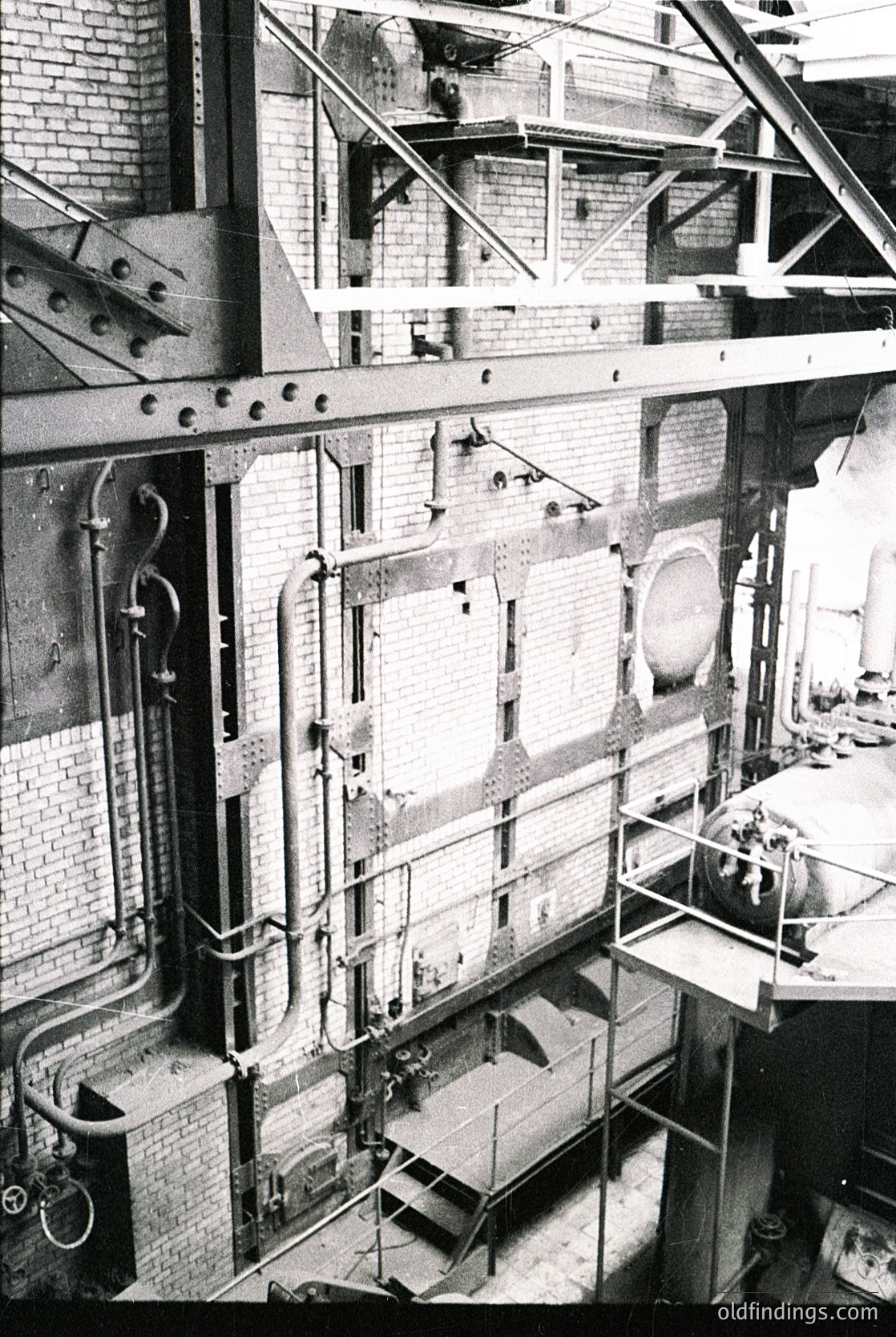 Industrial brick building interior with exposed steam pipes, scaffolding, and large circular/rectangular openings. Likely a 20th-century factory or power plant. Structural details suggest mid-century industrial architecture.