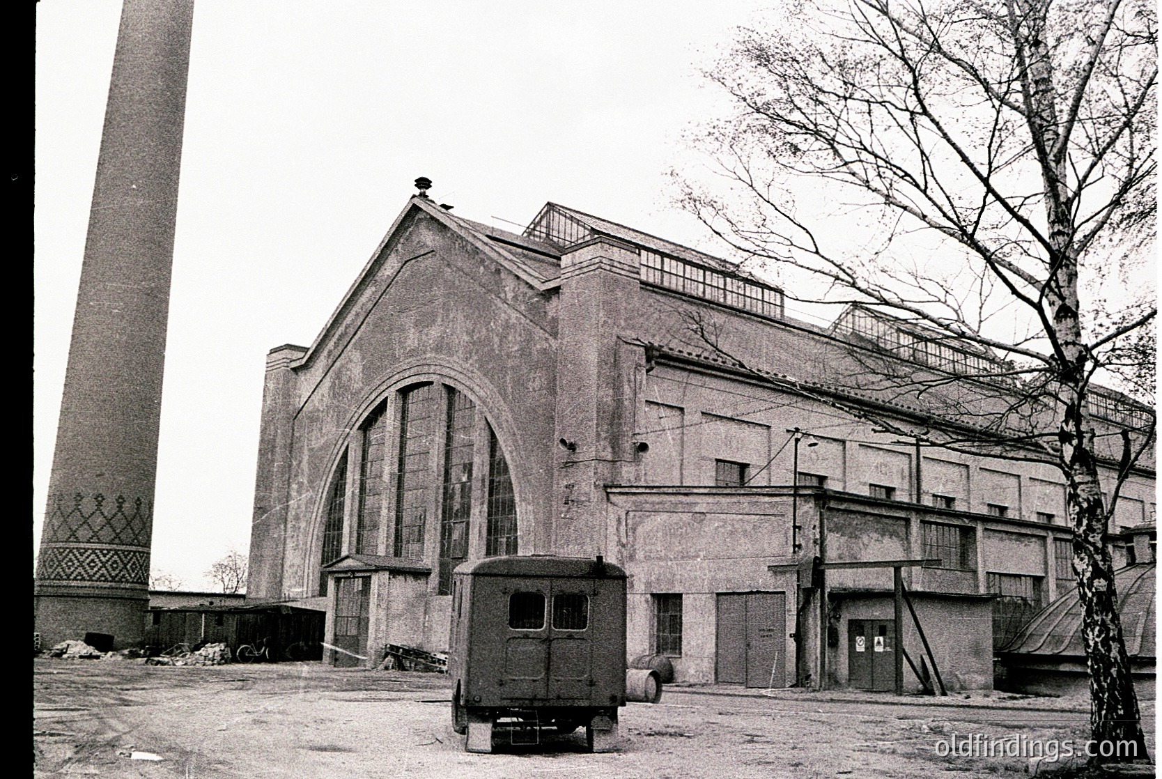 Mid-century industrial building with Brutalist concrete façade featuring large arched windows and minimalist detailing. A vintage utility truck parked in front suggests mid-20th-century setting. Adjacent smokestack hints at factory or power plant.