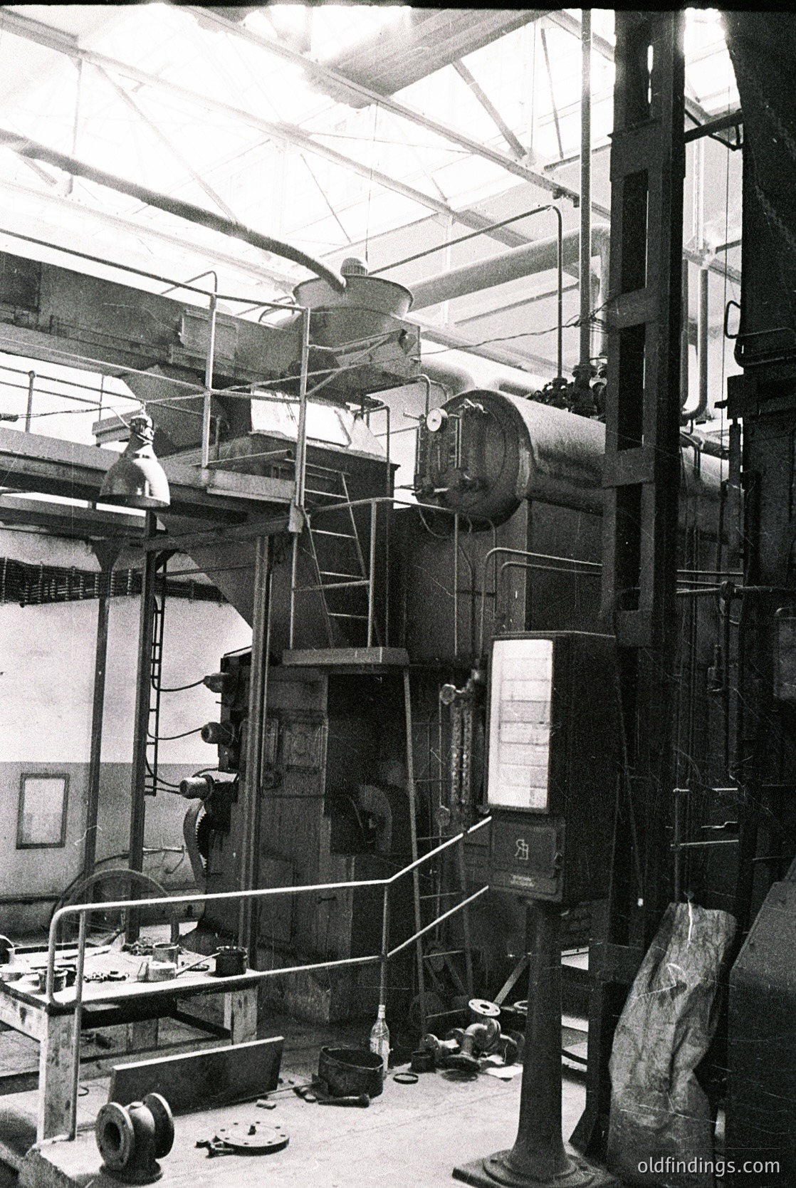 Industrial steam-powered machinery in a mid-20th century factory setting. Visible components include a large boiler, steam pipes, and a pulley system with exposed gears. The high ceiling and metal framework suggest a purpose-built facility for heavy manufacturing or textile production. The dated design reflects early-to-mid 1900s engineering.