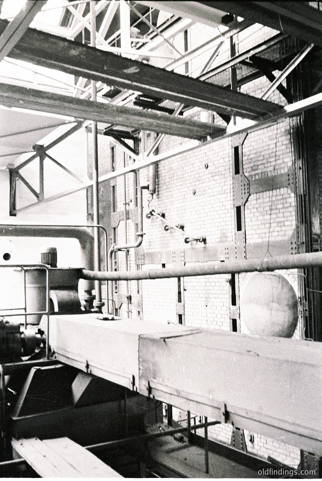 Industrial factory interior featuring exposed brick walls, steel beams, and conveyor systems. Mid-20th century machinery with pipes, valves, and a large cylindrical drum. Likely a manufacturing or processing plant.