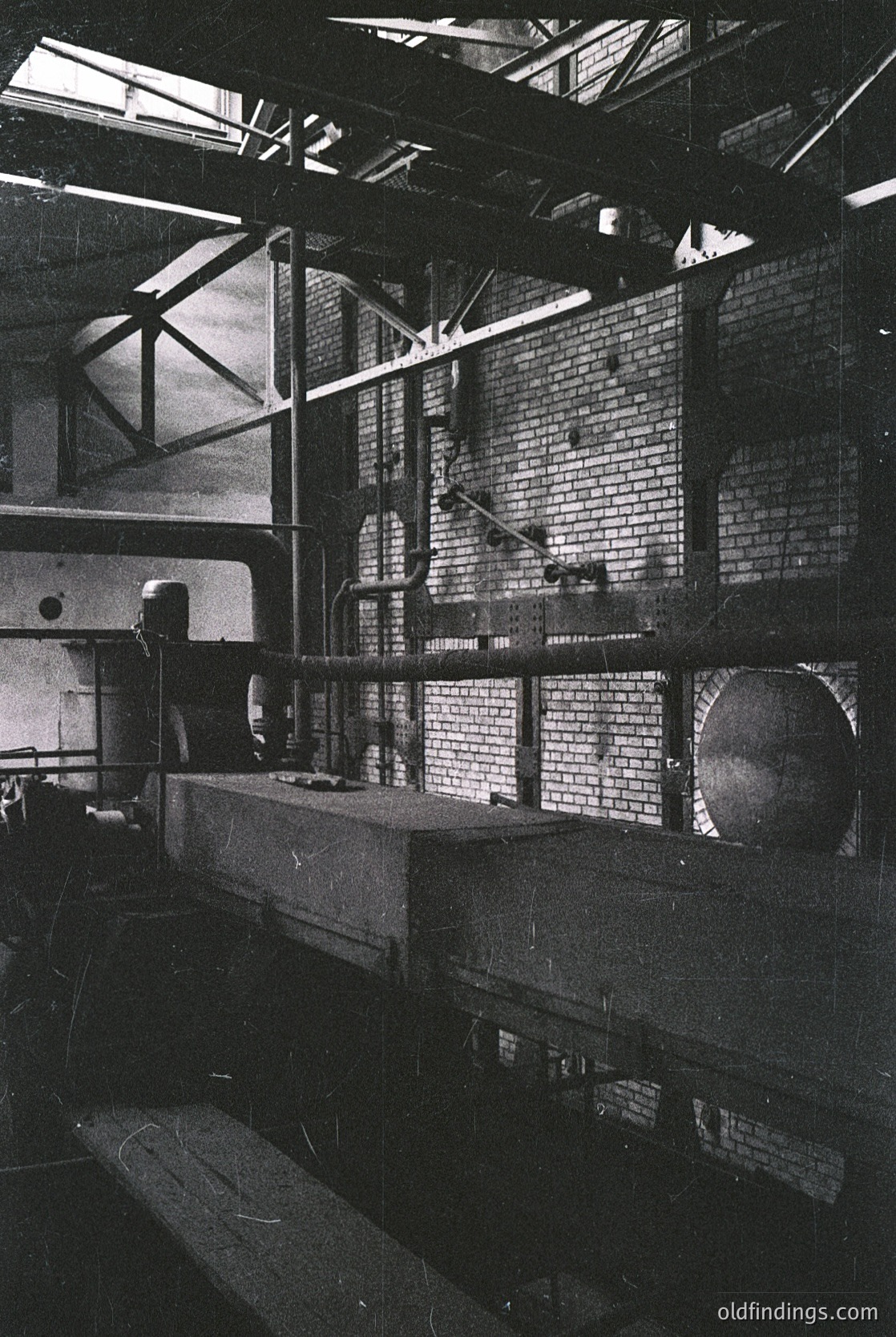 Industrial warehouse interior with exposed brick walls and high ceilings. Large metal beams and pipes dominate the space, suggesting mid-20th century manufacturing or storage. A long conveyor belt or platform runs horizontally, flanked by machinery and structural supports. Skylights allow natural light to filter through.