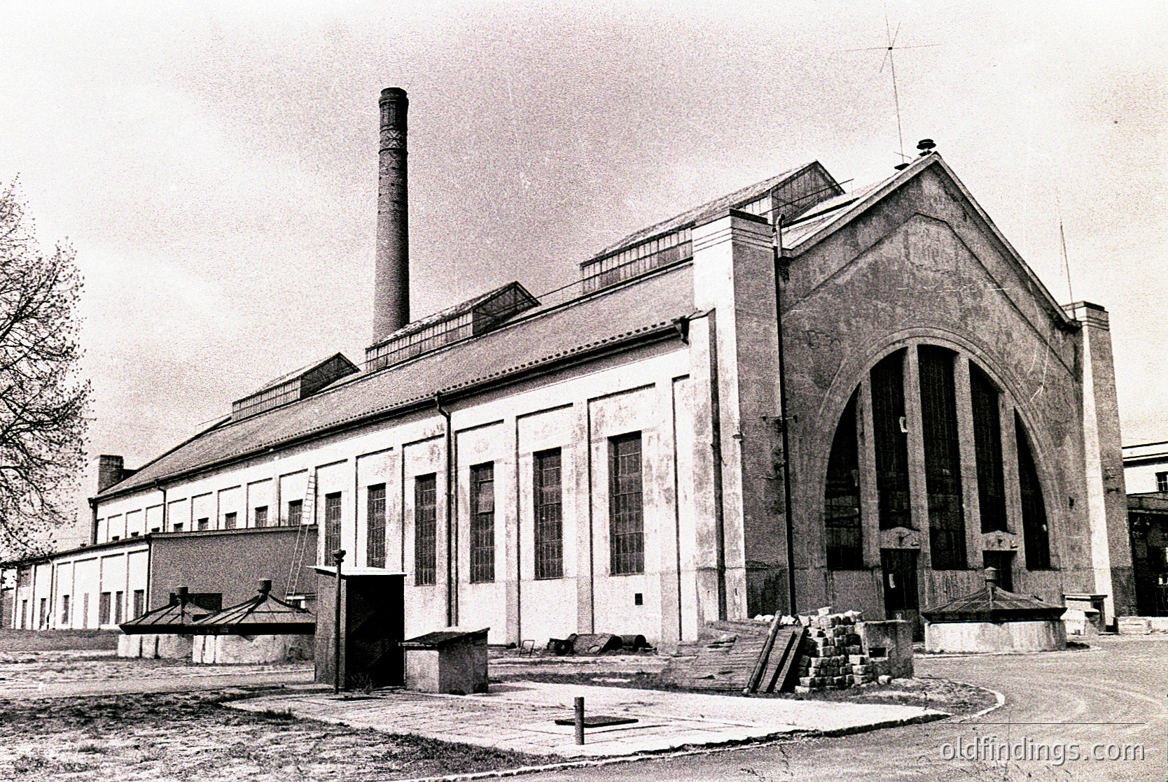 Industrial-era brick building with Art Deco-inspired arched entrance and tall chimney stack, likely a factory or power plant. Symmetrical facade with large rectangular windows and minimalist detailing. Overgrown vegetation suggests abandonment or disuse. Black-and-white, mid-20th century.