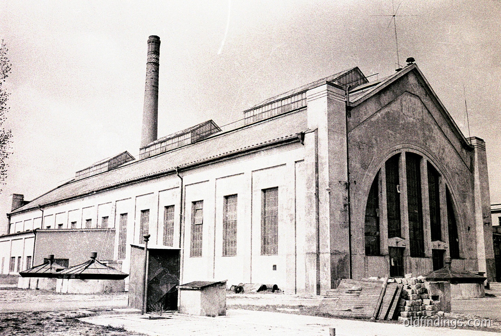 Industrial-era brick building with Art Deco-inspired arched entrance and tall chimney stack. Exterior shows signs of aging and weathering, with visible cracks and debris. Likely a factory or power plant from the early-to-mid 20th century.