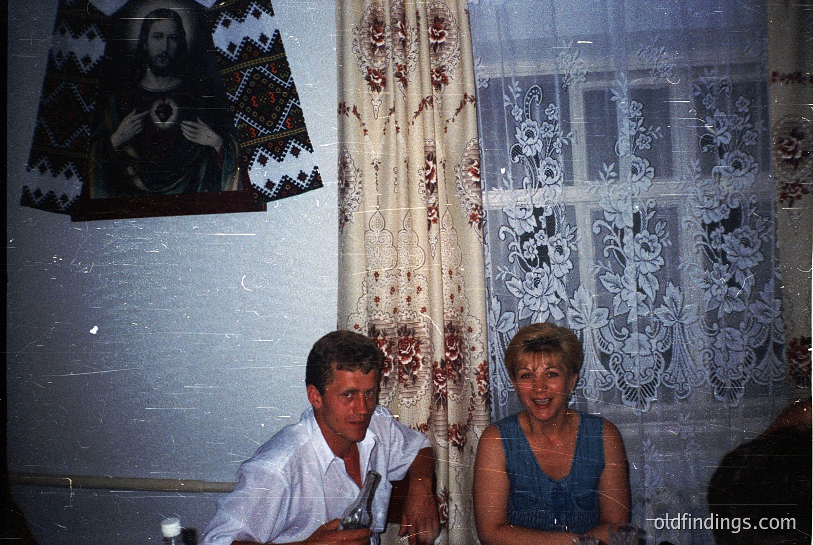 Vintage interior featuring two individuals seated at a table, framed by intricate lace curtains and a religious icon depicting Christ. The man wears a light-colored button-down shirt; the woman, a blue sleeveless top. Decor includes floral-patterned wallpaper and a framed embroidered textile. Likely Eastern European, mid-20th century.