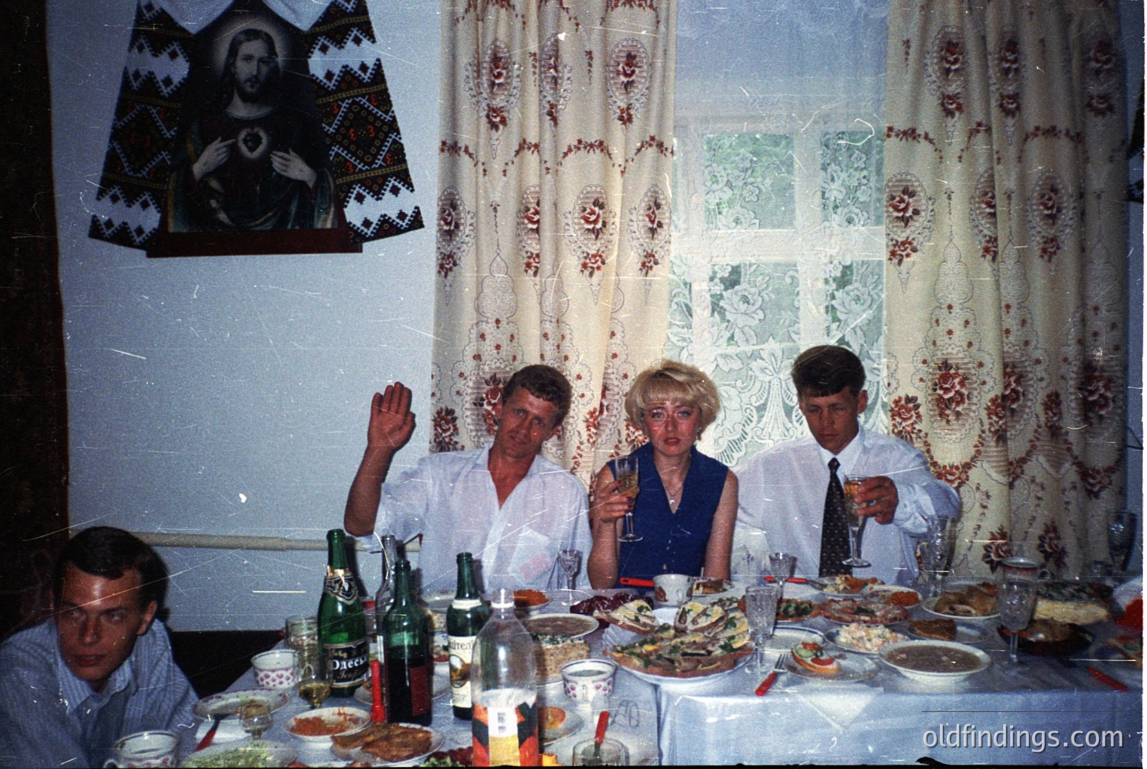 Vintage indoor gathering featuring four adults seated at a long table laden with seafood, bottles, and glasses. Decor includes floral curtains, a religious icon, and a framed poster. Likely Eastern European, 1970s-1980s.