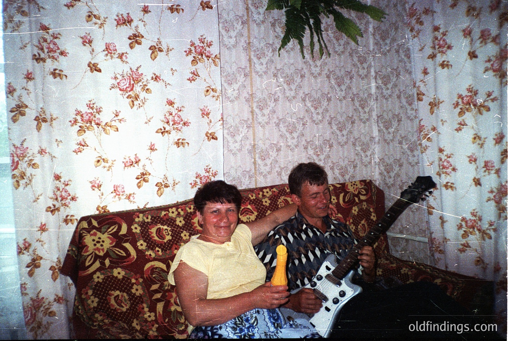 Vintage indoor portrait featuring two individuals in a patterned floral sofa setting, likely Eastern European 1980s-90s. Man plays electric guitar; woman holds a yellow object. Floral wallpaper and retro decor suggest mid-century domestic life.