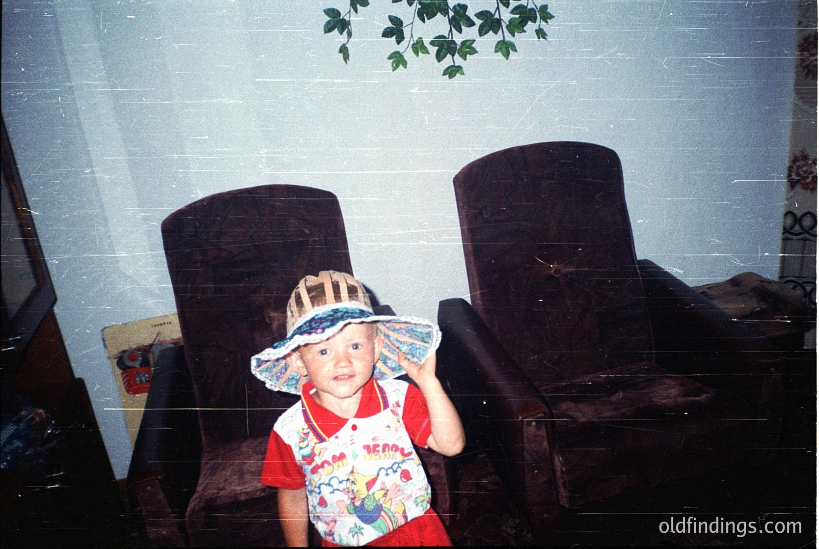 Vintage indoor portrait of a young child wearing a wide-brimmed hat with floral patterns and a colorful, cartoon-themed shirt. Dark wooden armchairs frame the scene, suggesting a mid-20th-century setting. Lighting and grain indicate a 1970s-1980s photo.