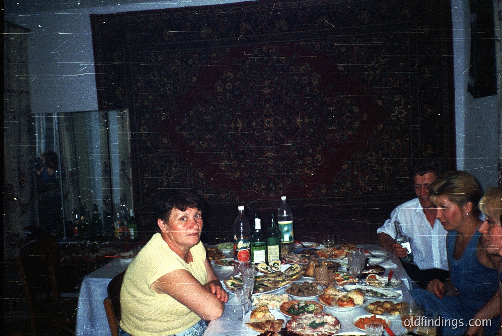 Vintage indoor gathering featuring a long dining table laden with seafood platters, bottles, and glasses. Three adults seated, one woman in a yellow sweater smiling at camera. Floral wallpaper and industrial-style lighting. Likely Eastern European dining culture, 1980s-1990s.