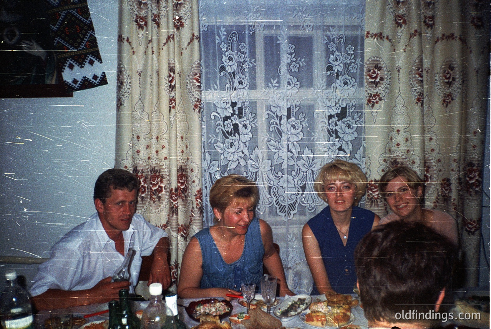 Vintage indoor gathering featuring four adults seated at a table with patterned lace curtains and floral wallpaper. Mid-century dining setup includes a bottle, plates with food, and a blurred camera flash. Likely 1970s–1980s Eastern European home setting.