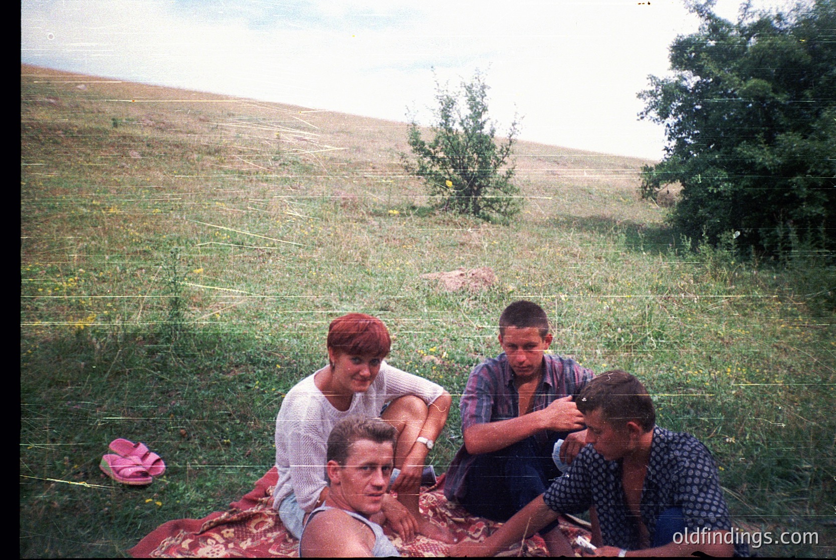Vintage outdoor portrait of three individuals seated on grassy hillside, likely 1960s-1970s. One person’s hair is being styled with a comb, suggesting a candid, informal moment. Faded colors and grainy texture indicate aged film. Rural or semi-rural setting with barbed wire fence and scattered trees.