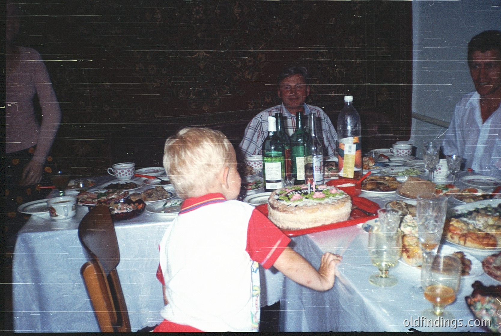 Vintage family gathering at a festive table, likely 1980s–1990s. A child in a red-and-white striped shirt holds a birthday cake with candles, surrounded by plates of food, wine bottles, and glasses. Dark wood furniture and patterned wallpaper suggest a mid-century European home. Warm, intimate setting with blurred motion indicating candid, lively moments.
