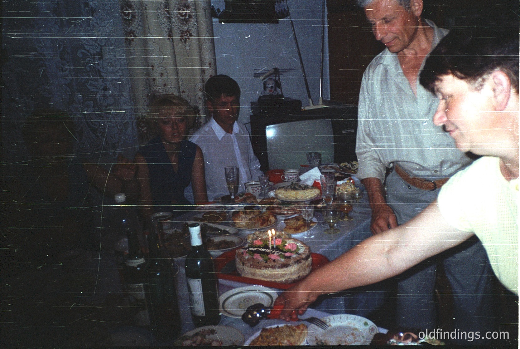 Family celebration with birthday cake featuring lit candle, likely 1980s-1990s indoor setting. Three adults and one child gather around a table adorned with plates, glasses, and traditional baked goods. Vintage refrigerator and patterned wallpaper suggest mid-century home decor.