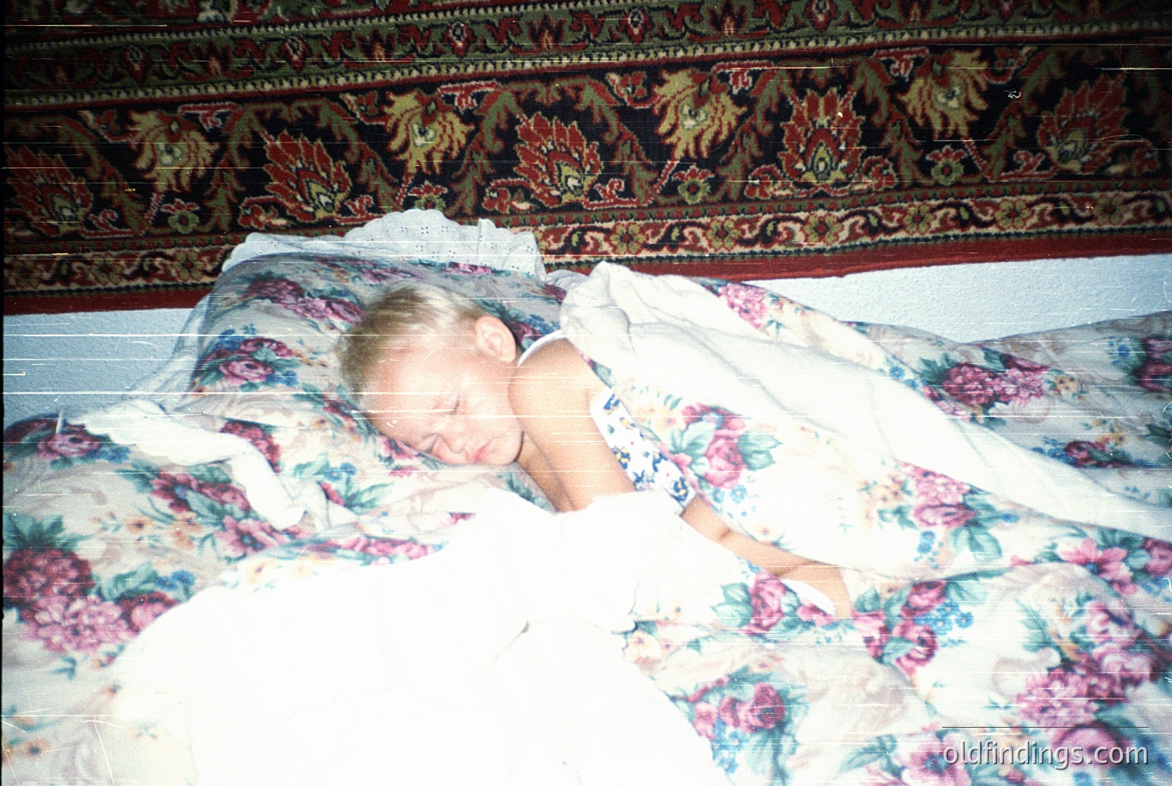 Child resting on floral-patterned bedding with ornate wallpaper featuring floral and animal motifs, likely mid-20th century interior. Warm, vintage color palette suggests 1960s–1980s domestic setting.