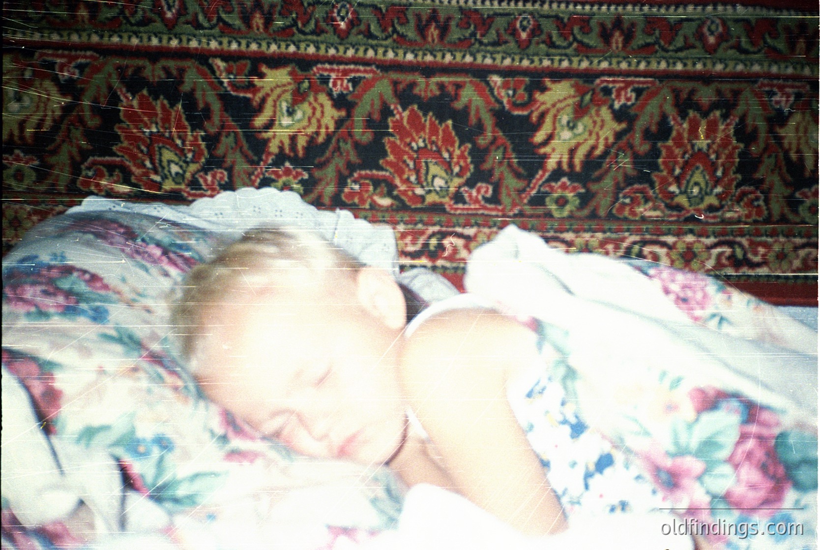 Vintage floral-patterned bedding with ornate red/gold wallpaper backdrop. Infant sleeping peacefully under a light blanket, likely mid-20th century. Warm, nostalgic family moment.