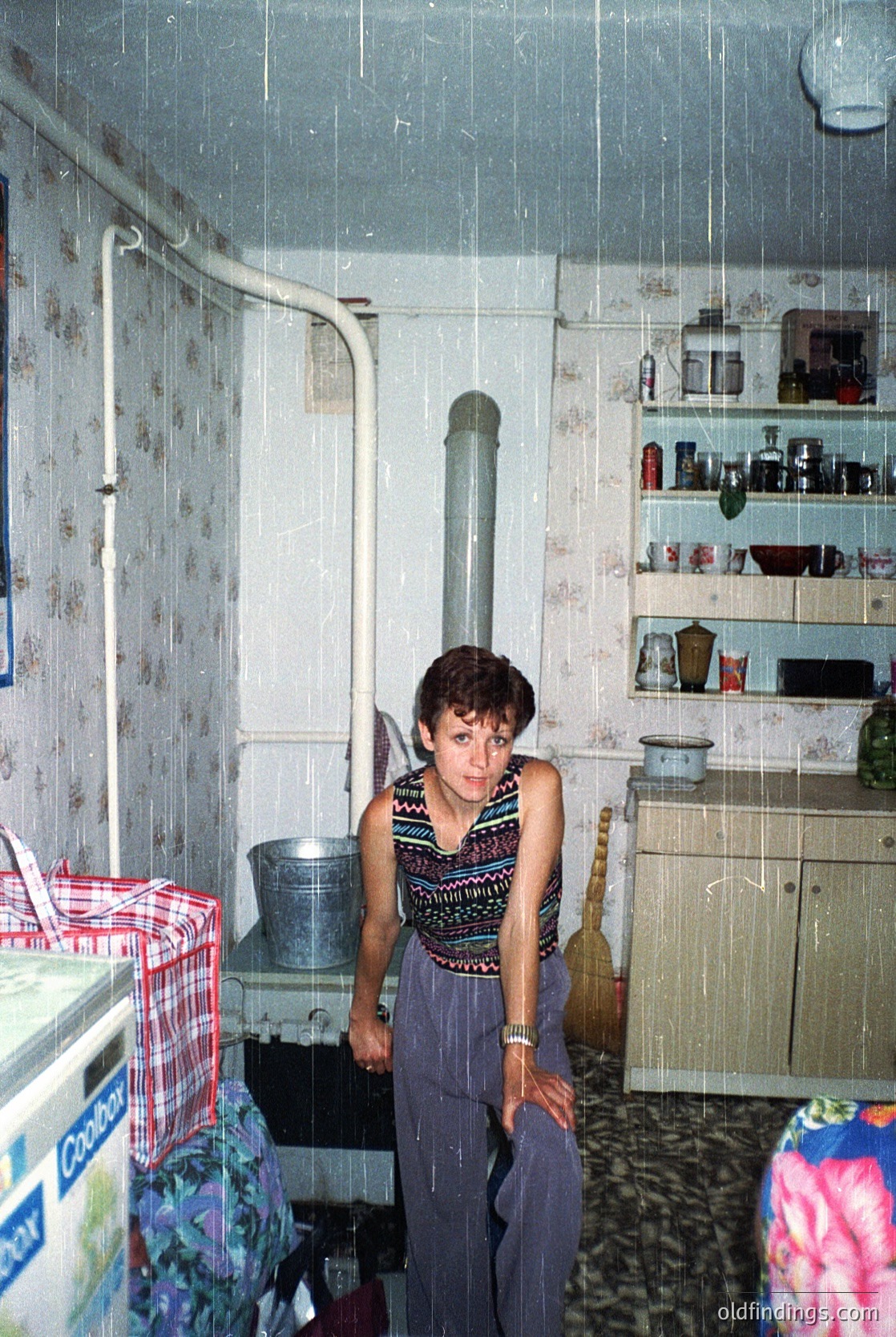 A woman in a patterned sleeveless top and wide-legged pants stands in a modest kitchen, leaning against a stove with a metal pot. Whitewashed walls, floral wallpaper, and a tiled floor reflect utilitarian Soviet-era design. Shelves hold mismatched jars, a stove pipe vents upward, and a refrigerator sits to the left. The scene suggests domestic life in Eastern Europe during the mid-20th century. [Mid-20th-century Soviet-style kitchen interior with woman standing beside stove ]