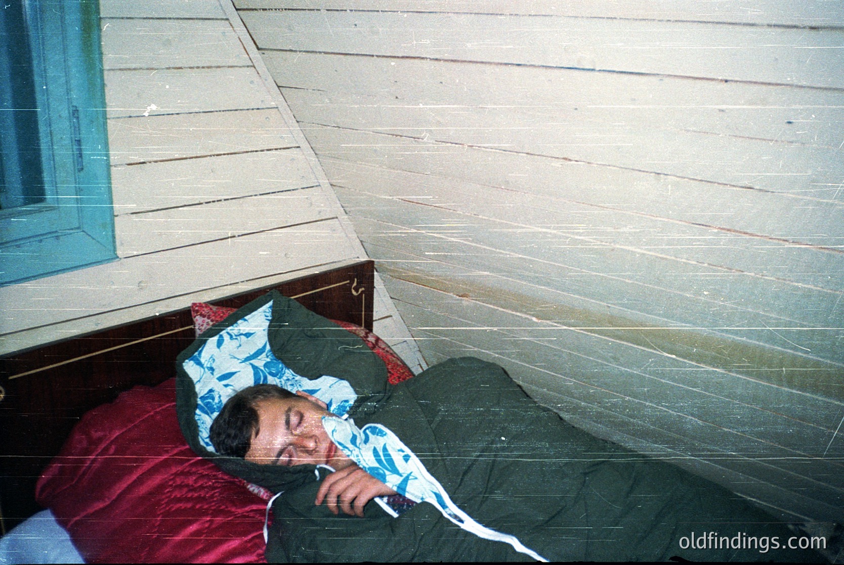 Vintage-style bunk bed with a single sleeper resting on the lower mattress, covered by a patterned blue pillowcase and olive-green blanket. Wooden paneling and a frosted glass window frame suggest a mid-20th century interior. Warm, nostalgic lighting enhances the retro aesthetic.