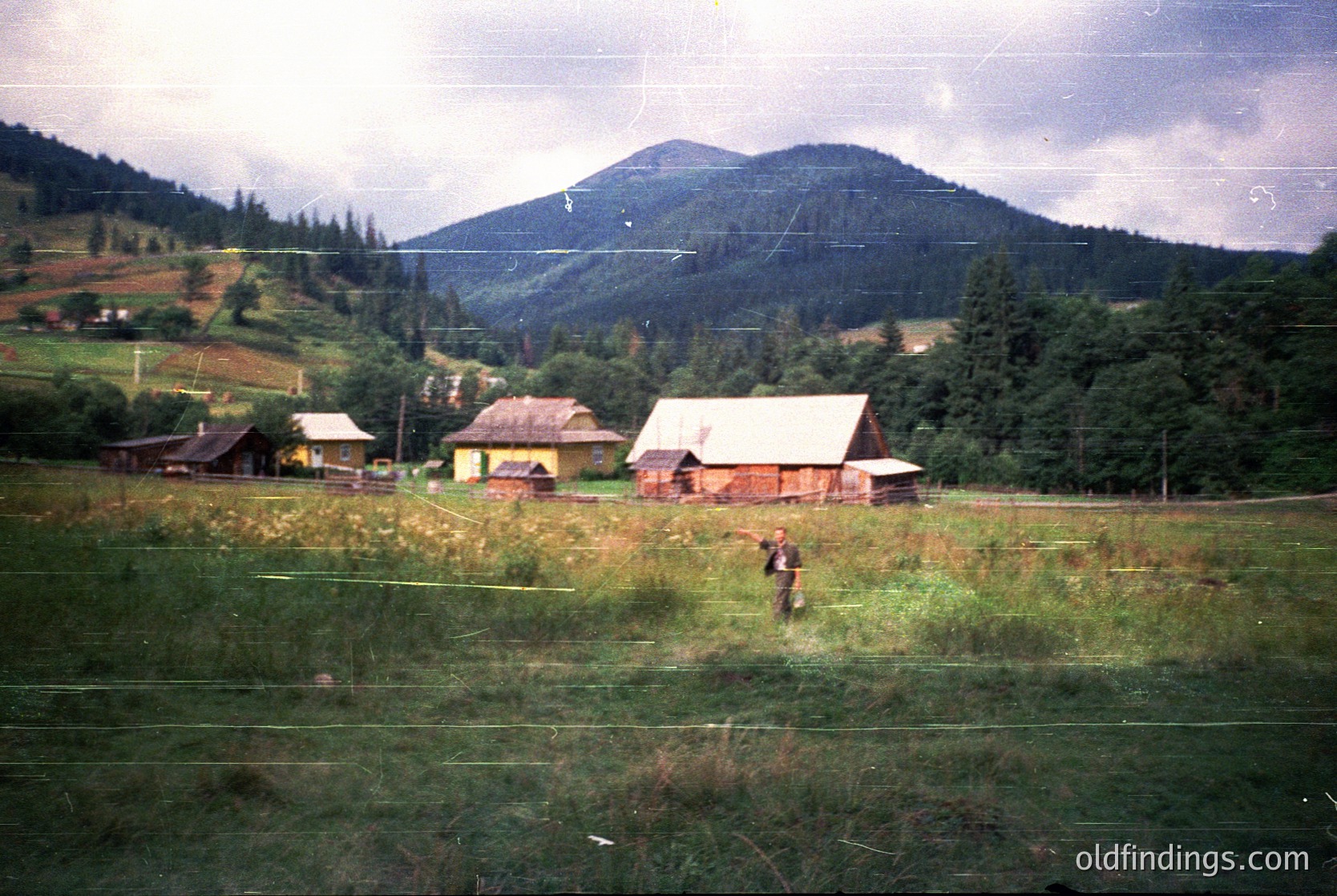 Vintage rural scene featuring rustic wooden farmhouses with pitched roofs, surrounded by lush green fields and forested hills. A lone figure in dark clothing walks near a fence, suggesting mid-20th century agricultural life. Overcast skies enhance the nostalgic atmosphere.