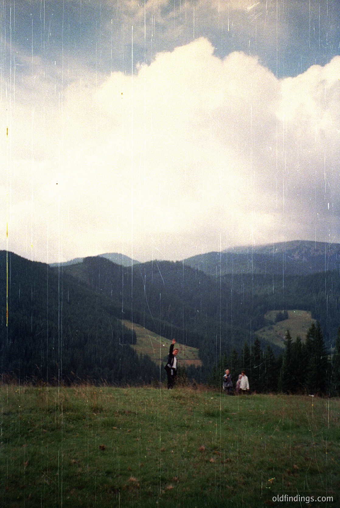 Three figures stand on a grassy hilltop under dramatic, rain-soaked skies, framed by vertical rain streaks. Lush forested valleys and rolling mountains stretch into the distance, suggesting a European alpine region. The composition captures a moment of solitude amid nature’s raw beauty.