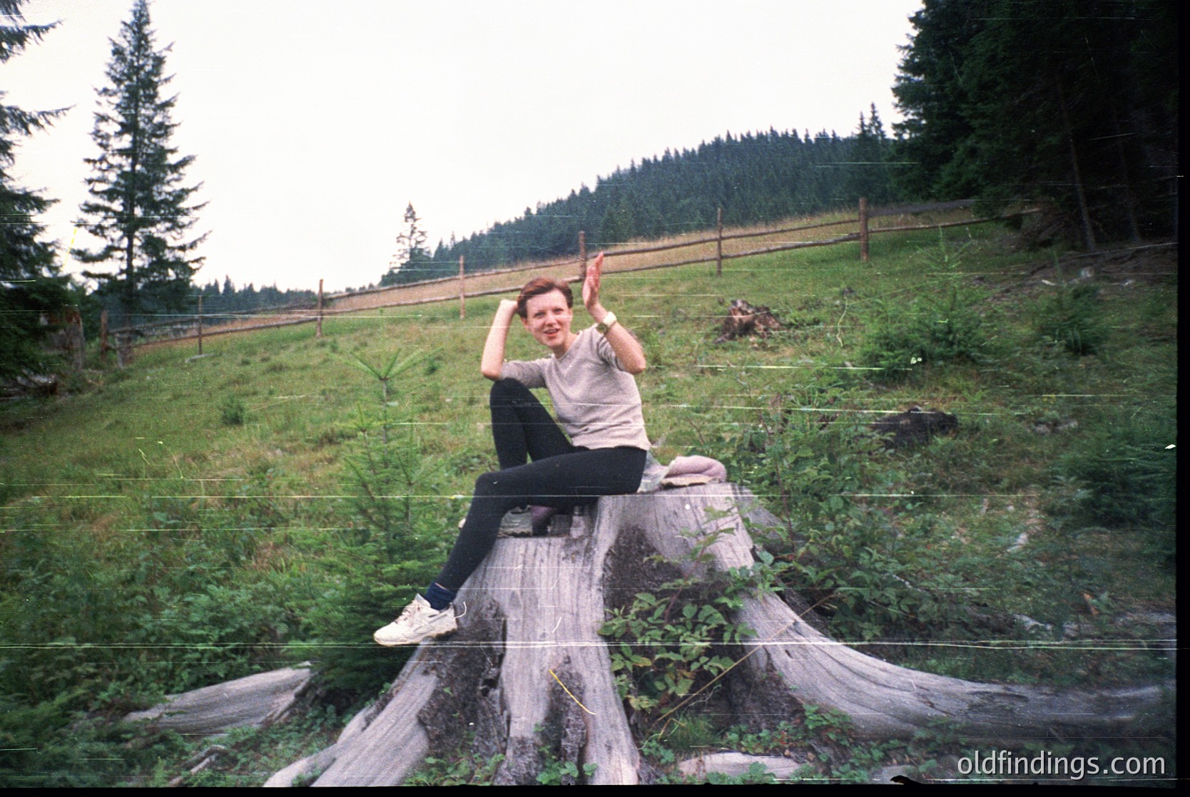 Vintage candid of person posing on a stump in a forested hillside, mid-1990s. Casual attire includes a light jacket, dark pants, and sneakers. Wooden fence and dense coniferous trees in background.