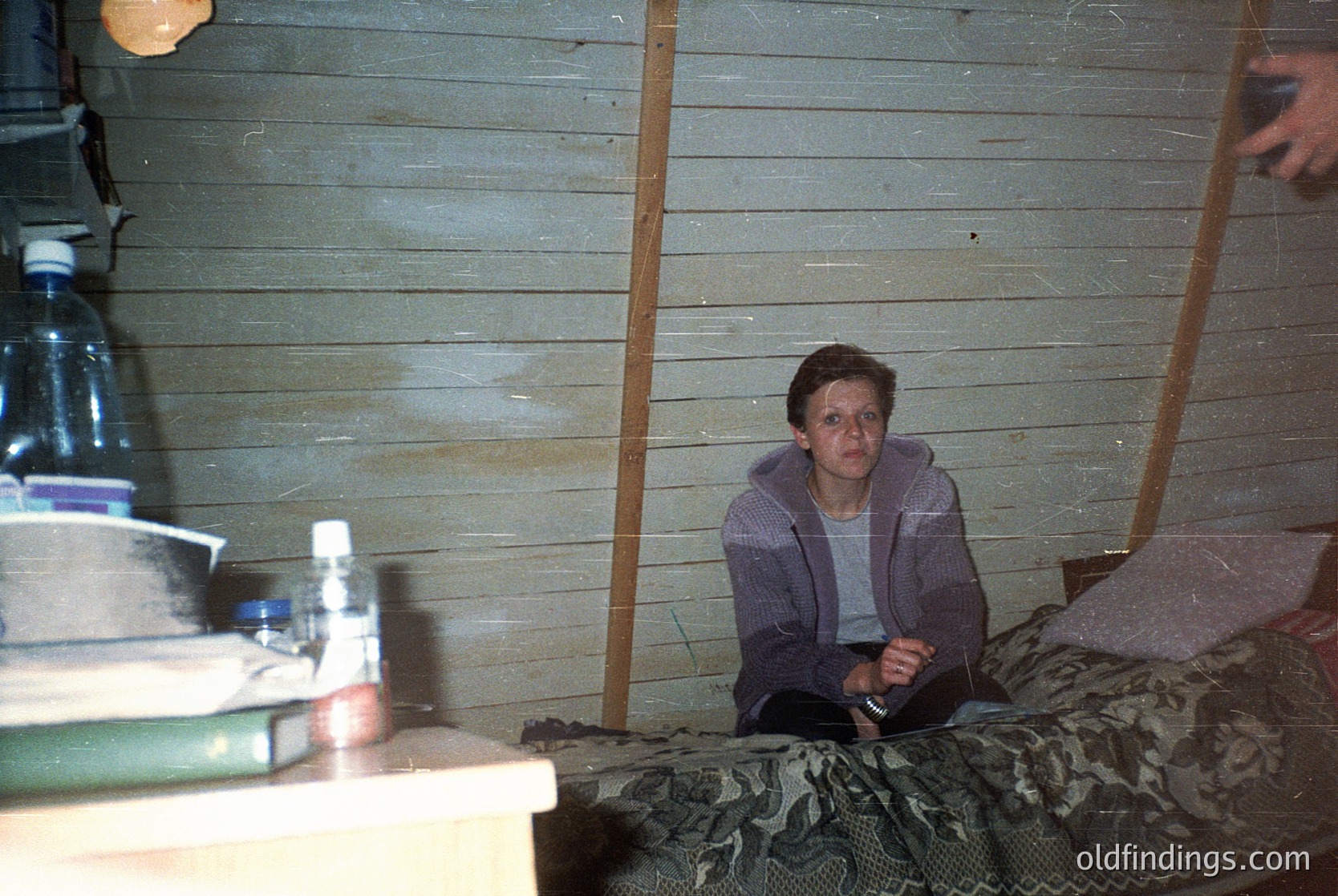 Indoor shot of a person seated on a camouflage-patterned bed in a rustic wooden cabin. Bottles of water and a plastic container on a shelf beside them. Warm lighting suggests evening or low natural light. Likely rural setting, mid-20th century.