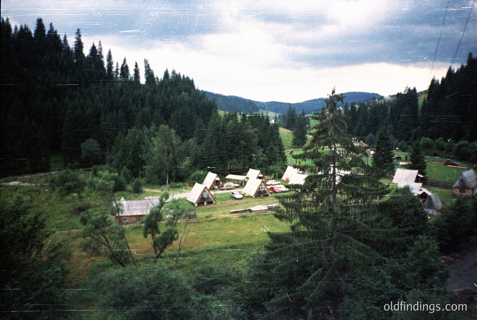 Aerial view of a rural campsite nestled in a forested valley, likely mid-20th century. Several pitched tents and a few small wooden cabins dot the grassy clearing surrounded by dense coniferous trees. Overcast skies and misty hills frame the scene, suggesting a mountainous or alpine region. The vintage color tone and power lines indicate historical documentation value.