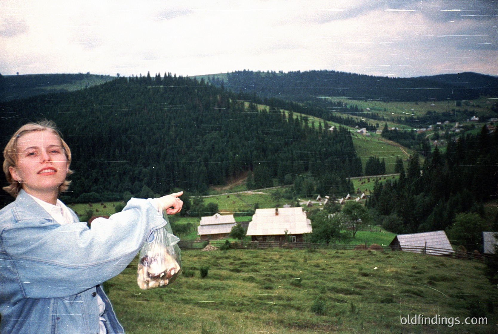A woman in a denim jacket and light blue shirt poses on a grassy hillside, pointing toward a valley with dense forest and scattered rural homes. She holds a mesh bag with small, round objects. Likely 1990s European countryside, possibly .